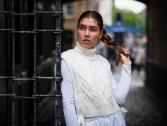 Woman in white turtleneck vest with braided hair standing by gate outdoors.