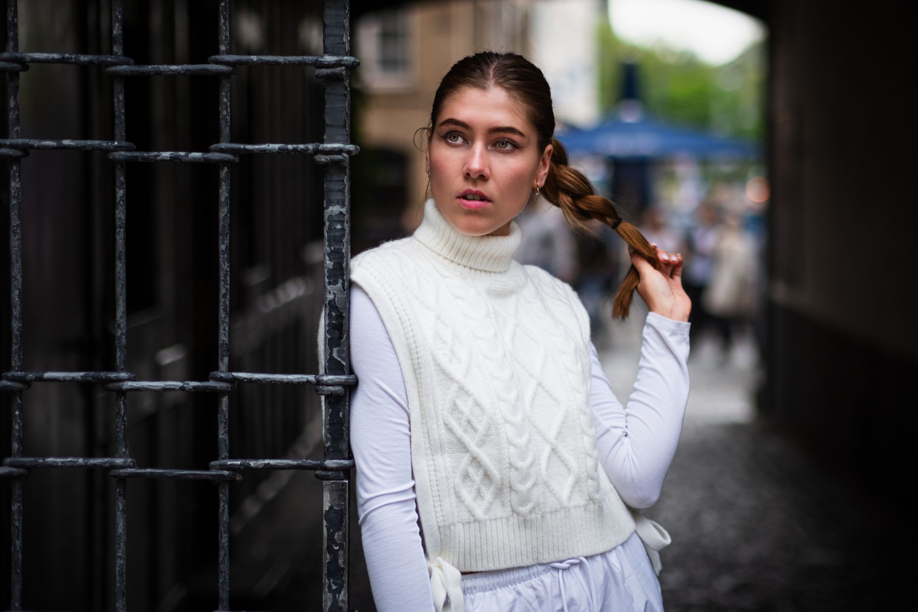 Woman in white turtleneck vest with braided hair standing by gate outdoors.